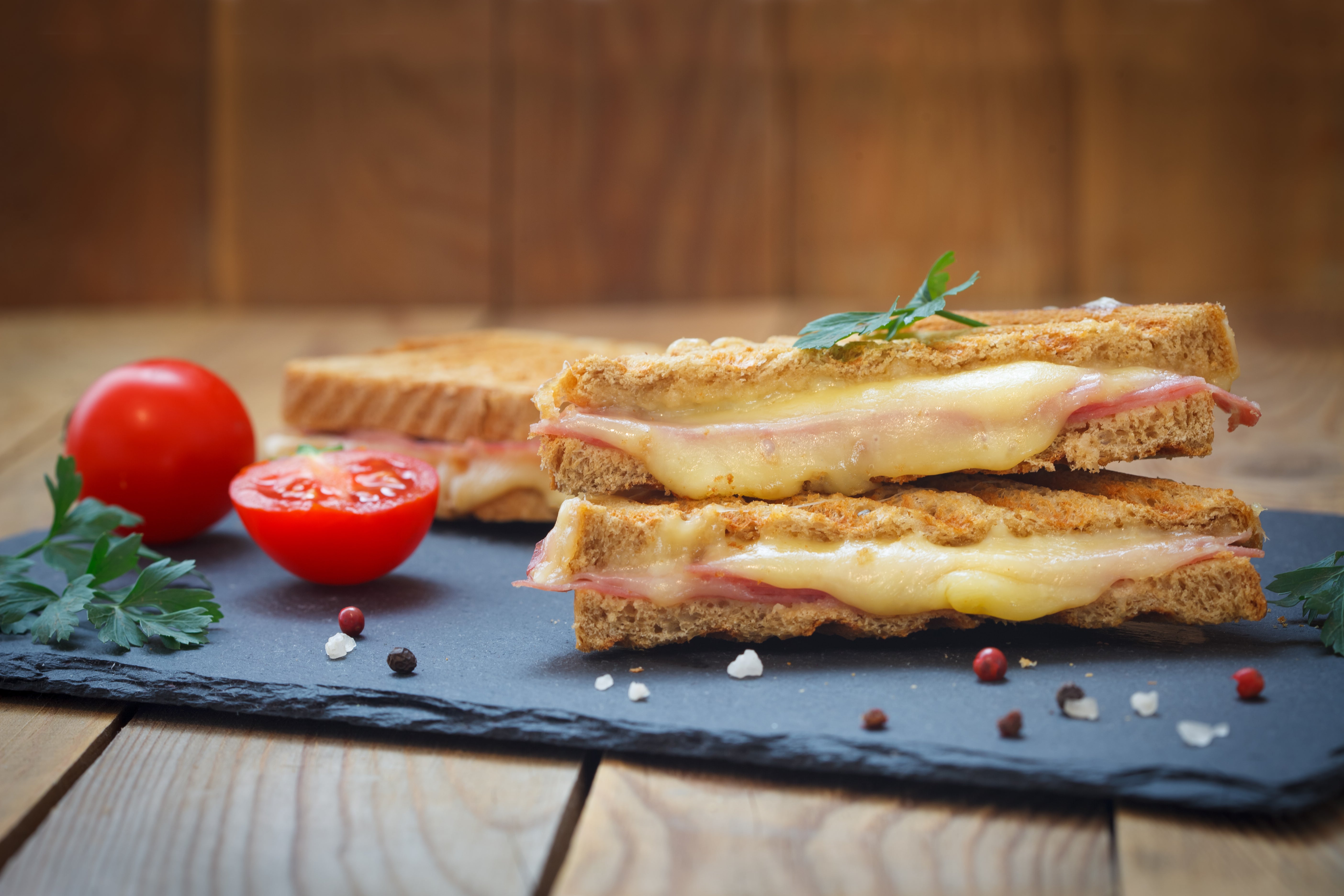 Three grilled cheese sandwiches on a slate board with tomatoes and herbs on a wooden background