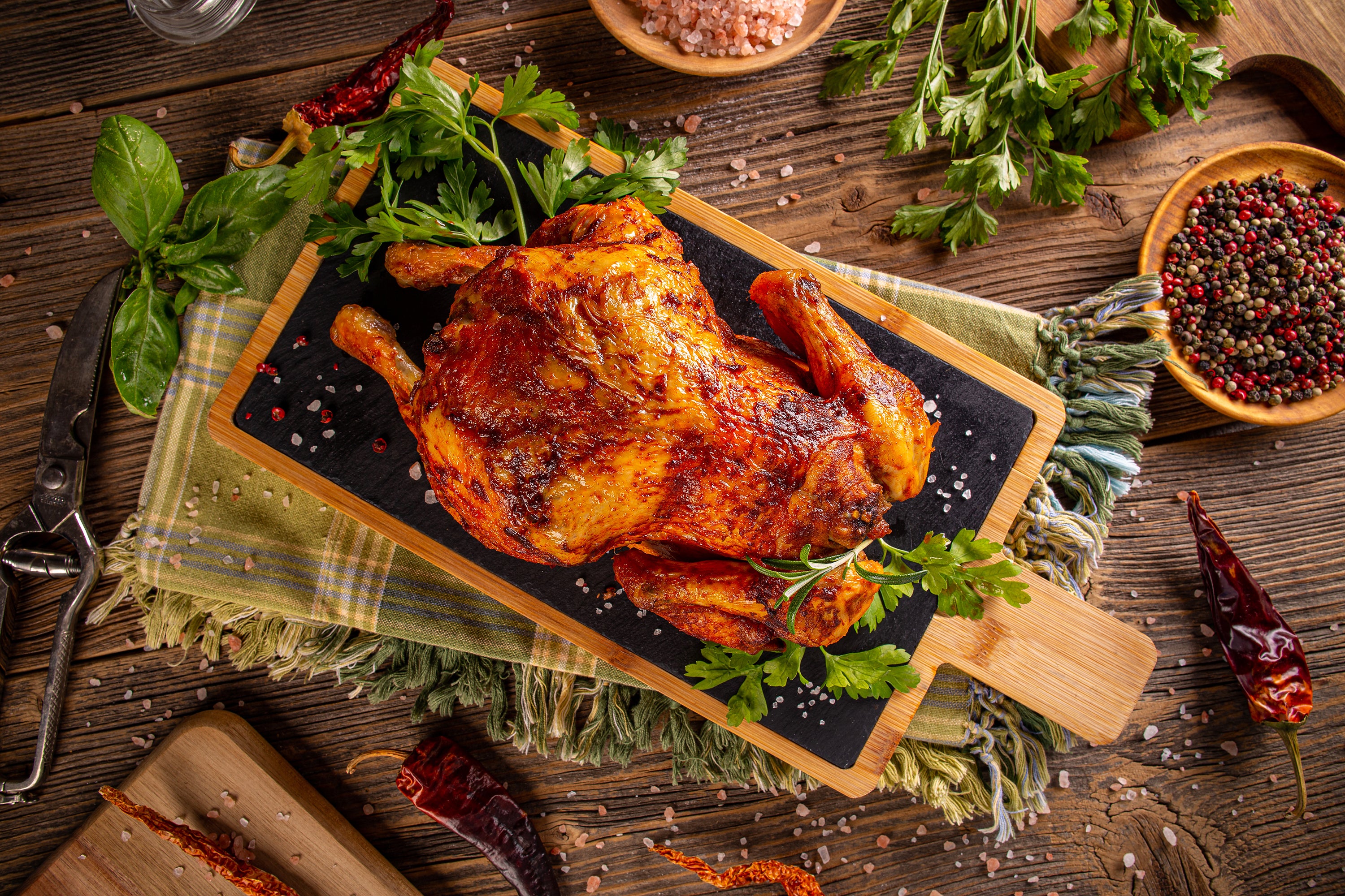 Roasted chicken on a wooden cutting board with herbs and spices on a rustic wooden table.